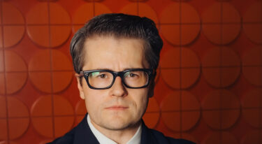 Portret moškega z očali pred oranžnim vzorčastim ozadjem.