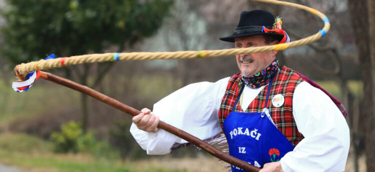 Fotografija od blizu prikazuje nastopajočega v folklorni/pustni opravi: črn klobuk z okrasjem, karirast telovnik čez belo srajco in moder predpasnik z napisom »POKAČ«. Z desnico vihti dolg pleten bič z barvnimi trakovi; bič opisuje lok pred njegovim obrazom. Ozadje je zunanje, rahlo zamegljeno (drevesa, trava).