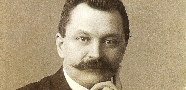 Črno-beli portret moškega z brki v obleki; roka je prislonjena k obrazu.