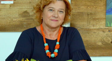 Portret ženske z oranžno-turkizno ogrlico; v ospredju so sončnice, v ozadju lesena stena.