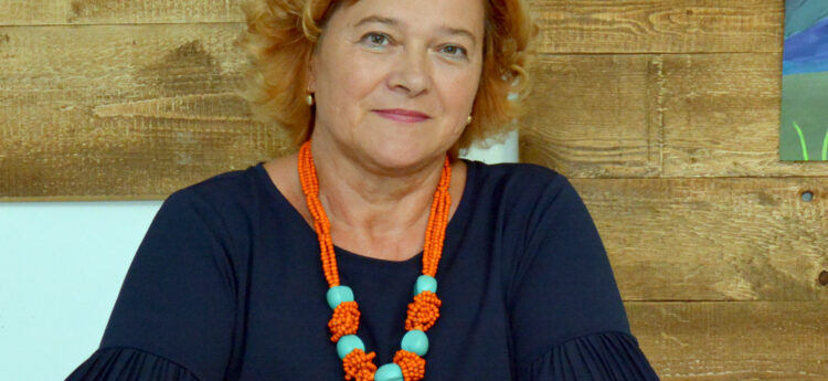 Portret ženske z oranžno-turkizno ogrlico; v ospredju so sončnice, v ozadju lesena stena.