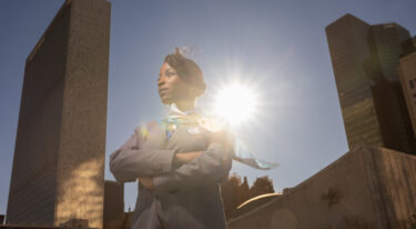 Ženska v uniformi stoji pred visokimi stavbami, osvetljena od zadaj s sončnim žarkom.