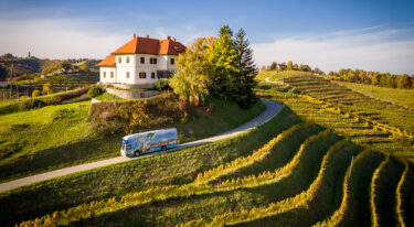 Bibliobus vozi po vijugasti cesti med vinogradi; v ozadju je bela hiša z rdečo streho na griču.