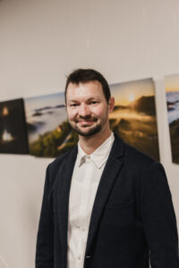 Portret avtorja v temnem suknjiču in beli srajci pred razstavljenimi fotografijami.